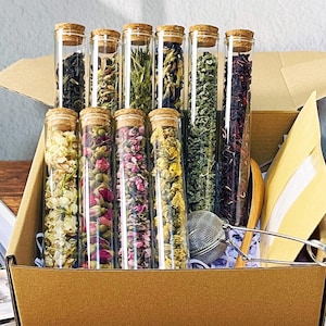 May include: A gift set of assorted teas in clear glass tubes with cork stoppers, arranged in a cardboard box. The tubes contain various dried herbs and flowers, with a stainless steel tea infuser. The box is open, revealing the contents.
