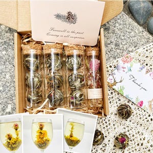 May include: A gift box filled with six glass tubes containing blooming tea flowers. The tubes are arranged in two rows of three. The box is made of wood and has a brown finish. The box also includes a card with the text "Farewell is the past, coming is all surprises." and a small card that says "Thank you".