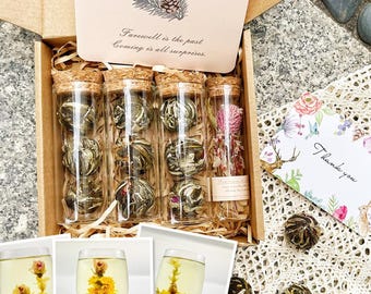 May include: A gift box filled with six glass tubes containing blooming tea flowers. The tubes are arranged in two rows of three. The box is made of wood and has a brown finish. The box also includes a card with the text "Farewell is the past, coming is all surprises." and a small card that says "Thank you".