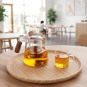 May include: A glass teapot with a wooden handle and infuser, filled with amber liquid, sits on a wooden tray. A matching glass teacup with tea is next to the teapot. The scene is set in a bright, airy room.