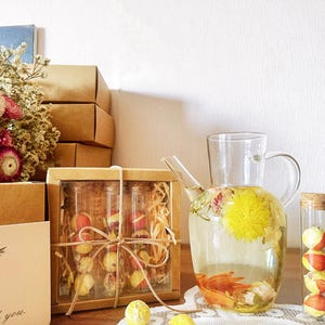 May include: A set of four glass vials filled with dried flowers and fruit, packaged in a brown box with a twine bow. A glass pitcher filled with water and dried flowers sits on a white tablecloth next to a vial with dried fruit.