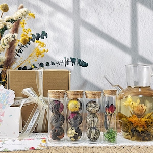 May include: Several glass bottles with cork stoppers, filled with various blooming tea varieties. A glass teapot with a blooming tea flower is also visible. A gift box with a "Happy Birthday" ribbon is in the background, along with dried flowers.