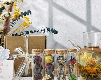 May include: A collection of glass tea infuser bottles with cork stoppers, each containing a different type of blooming tea. A glass teapot with a blooming tea flower is also present. A gift box with a ribbon that says "Happy Birthday" is in the background.