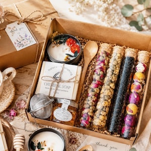 May include: A gift box filled with a variety of items, including a candle, soap, honey, tea tubes, and a wooden spoon. The box is brown and tied with twine. The soap is labeled "Oatmeal Soap". A tag reads "thank you".