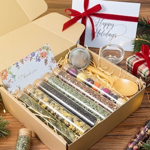 May include: A gift box with a variety of teas in glass tubes, a wooden spoon, and a tea infuser. The box includes a card that says "Thank you" and a card that says "Happy Holidays". The box is tied with a red ribbon.