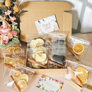 Może przedstawiać: Pudełko upominkowe wypełnione suszonymi plasterkami jabłek i pomarańczy, herbatą i laskami cynamonu. Artykuły są zapakowane w przezroczyste plastikowe torebki i ułożone w brązowym kartonowym pudełku. Dołączona jest karta z napisem "dziękuję!".