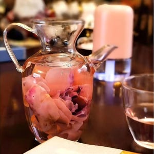 May include: A clear glass pitcher filled with pink liquid and pink flower petals. The pitcher has a handle and a spout. A glass of water and a pink cylindrical object are in the background.
