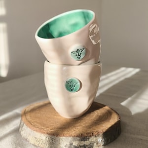 Op de afbeelding: Twee gestapelde keramische bekers, lichtroze met een getextureerde afwerking en een teal embleem. De binnenkant van de bovenste beker is teal. Ze staan op een houten onderzetter. Zonlicht creëert schaduwen.