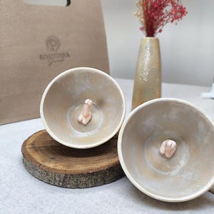 Puede incluir: Dos cuencos de cerámica beige con un diseño fálico en el centro, expuestos sobre una rodaja de madera. Una bolsa de papel marrón con el texto "ECHOTERRA Gourmet" está en el fondo, junto con un jarrón de flores secas.