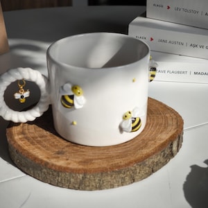 Puede incluir: Taza de cerámica blanca con un diseño de abeja. La taza tiene un asa con un pequeño amuleto de abeja. La taza está sobre un posavasos de madera.