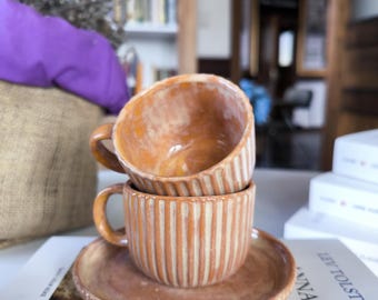 Puede incluir: Dos tazas de café de cerámica apiladas con un patrón a rayas en tonos marrones y beige. Las tazas están sobre un platillo con un patrón a rayas a juego. El platillo está sobre un libro titulado "ANNA KARENINA" de LEO TOLSTOY.