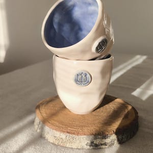 Op de afbeelding: Twee handgemaakte keramische bekers gestapeld op een houten onderzetter. De bekers zijn lichtroze met een blauw interieur en een blauw embleem. De onderzetter is een natuurlijke houten schijf met een schorsrand.
