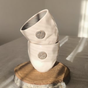 May include: Two off-white ceramic cups with a textured surface and a small, circular emblem. The cups are stacked on a round, wooden coaster. The cups have a unique, handmade appearance.
