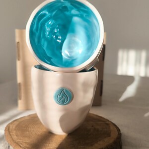 May include: Two ceramic cups, one stacked atop the other. The top cup has a bright turquoise interior, while the bottom cup is a soft pink with a turquoise emblem. The cups are on a wooden coaster.