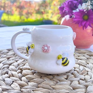 May include: White ceramic mug with a curved handle, adorned with three-dimensional bees and pink flowers. The mug sits on a woven mat, with a blurred background of flowers and foliage.