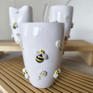 Puede incluir: Tazas de cerámica blanca decoradas con abejas y margaritas en 3D. Cada taza tiene una pajita de cristal transparente. Las tazas están sobre una bandeja de madera. Las abejas tienen rayas amarillas y negras, y las margaritas tienen centros amarillos.
