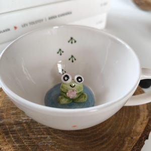 Puede incluir: Una taza de té de cerámica blanca con una rana verde que sostiene una flor rosa en su interior. La rana tiene ojos negros y una sonrisa blanca. La taza tiene huellas verdes en el interior.