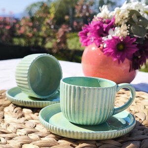 May include: Two aqua-coloured ceramic teacups and saucers with vertical ribbed detailing. The cups sit on a woven mat, with a blurred background of flowers and greenery. The teacups have a handle and are placed on matching saucers.