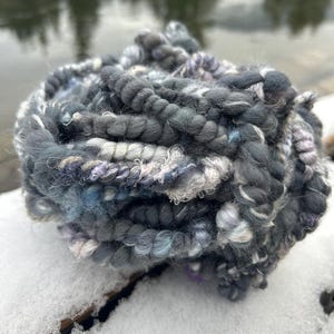 Puede incluir: Una madeja de hilo hilado a mano en tonos grises, azules y blancos. El hilo tiene un aspecto grueso y texturizado, con torsiones visibles y variaciones de color. El hilo está sobre una superficie nevada, con un fondo borroso de agua y árboles.