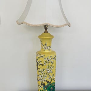 May include: A yellow ceramic table lamp with a white lampshade. The lamp base features a floral design with white blossoms and green leaves.
