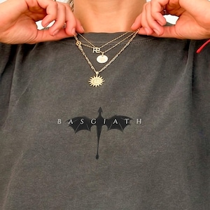 May include: Dark grey t-shirt featuring a dragon graphic and the word "BASGIATH" underneath. The shirt has a faded appearance. The model is wearing gold necklaces.