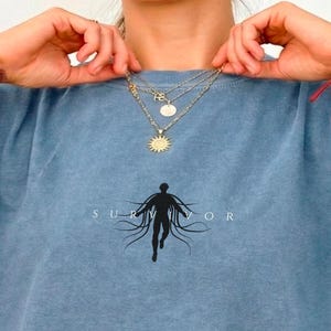 Pode incluir: Uma camiseta azul com a palavra "SURVIVOR" e um gráfico de uma pessoa com linhas fluidas. A camiseta é de cor azul lisa e tem um corte folgado. A pessoa na imagem está usando colares dourados.