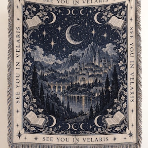 May include: A rectangular woven throw blanket featuring a dark blue and cream scene of a mountain city by a lake under a crescent moon and stars. The border displays floral vines, open books, and the repeated text SEE YOU IN VELARIS.