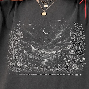 Puede incluir: Una camiseta gris grafito con un estampado gráfico blanco de un paisaje montañoso bajo una luna creciente y un cielo estrellado, enmarcado por flores silvestres. El texto dice: TO THE STARS WHO LISTEN AND THE DREAMS THAT ARE ANSWERED.