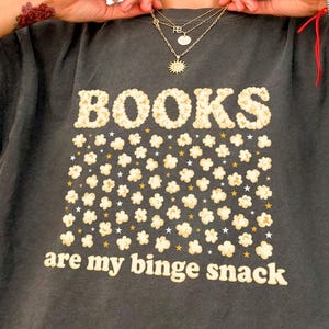 May include: Dark gray t-shirt with the words "BOOKS" and "are my binge snack" in a popcorn-like font. The design includes popcorn and star graphics. The shirt is a casual, comfortable style.