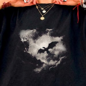 May include: Black t-shirt featuring a dragon silhouette against a cloudy background. The dragon is in flight, with its wings spread wide. The shirt is a dark color and has a classic crew neck design. The image is a graphic print.