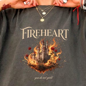 May include: A charcoal gray t-shirt featuring a graphic print of a castle surrounded by orange flames and red roses. The text FIREHEART is printed in a cream serif font above the castle, with the text you do not yield printed below it.