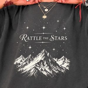 Może przedstawiać: Grafitowy t-shirt z białym nadrukiem graficznym przedstawiającym pasmo górskie pod rozgwieżdżonym niebem. Napis RATTLE THE STARS jest nadrukowany stylizowaną białą czcionką nad górami, w otoczeniu małych czteroramiennych ikon gwiazd.