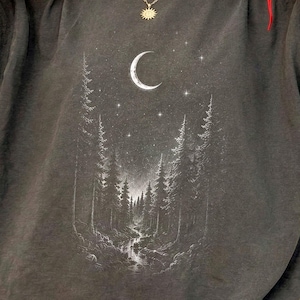 May include: A charcoal gray short-sleeve t-shirt featuring a white graphic print of a forest landscape at night. The design includes tall pine trees, a flowing stream, a crescent moon, and scattered stars centered on the front of the garment.