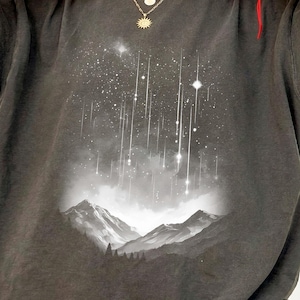Könnte beinhalten: Dunkelgraues T-Shirt mit einem Bergmotiv. Der Aufdruck zeigt einen Sternenhimmel mit fallenden Lichtern über einer Bergkette. Das Shirt hat eine lockere Passform und eine Vintage-Waschung.