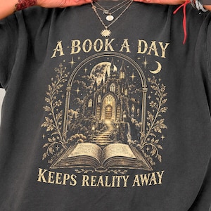 May include: A graphite-coloured t-shirt featuring a gold-toned graphic print of an open book with a castle rising from its pages. The text A BOOK A DAY KEEPS REALITY AWAY is printed in a serif font above and below the central illustration.