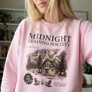 May include: A light pink sweatshirt with the text "MIDNIGHT READING SOCIETY" in dark brown. Below the text is a graphic of a cabin in the woods with mountains and trees. The sweatshirt also has the text "Candles & Secrets" and "Just bring your cozy pajamas."