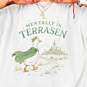 May include: A white t-shirt featuring a graphic of a white duck wearing a gold crown and a green cape. The text "MENTALLY IN TERRASEN" is printed above the duck, which is walking toward a detailed illustration of a castle with mountains in the background.