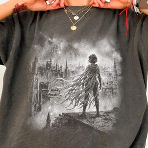 Puede incluir: Camiseta gris oscuro con una ilustración en blanco y negro de una figura con capa mirando una ciudad gótica. La imagen es detallada e impactante. La camiseta tiene un aspecto vintage.