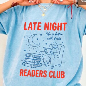 以下が含まれることがあります： 「LATE NIGHT READERS CLUB」の赤い文字が入った水色のTシャツ。三日月、星、椅子で本を読む人のイラストが描かれています。「life is better with books」の文字も含まれています。