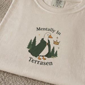 May include: An off-white short-sleeve t-shirt featuring a graphic of a white duck wearing a dark green cloak and a small gold crown. The design includes dark green pine trees and the text "Mentally In Terrasen" in a dark green font.