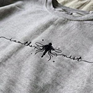 以下が含まれることがあります： 「i am the mist」の刺繍が施されたヘザーグレーのスウェットシャツ。テキストの下には、触手と星を持つ人物の黒いグラフィックがあります。スウェットシャツは柔らかい素材で作られています。