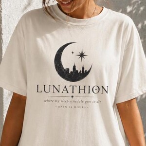 以下が含まれることがあります： オフホワイトの半袖Tシャツ。黒い三日月のグラフィックの中に街のスカイラインと星が描かれています。中央には「LUNATHION, where my sleep schedule goes to die, OPEN 24 HOURS」という文字が濃い色のフォントでプリントされています。