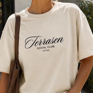 May include: Cream-colored t-shirt with the text "Terrasen Social Club" in a stylish script. The shirt has short sleeves and a relaxed fit. The model is wearing blue jeans and a brown tote bag.