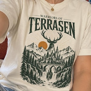 May include: An off-white short-sleeve t-shirt featuring a dark green graphic print. The design displays the text "WARRIORS OF TERRASEN" above an illustration of a stag, a mountain range, a sun, a castle on a bridge, and a waterfall surrounded by trees.