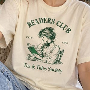 May include: A cream-coloured short-sleeve t-shirt featuring a dark green graphic of a person reading a book with a teacup. The text reads READERS CLUB, ESTD 1888, and Tea & Tales Society. The design is centred on the chest of the garment.