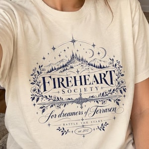Puede incluir: Una camiseta de manga corta de color crema con un estampado gráfico azul oscuro. El diseño incluye un paisaje de montaña, una espada, detalles florales y el texto "FIREHEART SOCIETY for dreamers of Terrasen RATTLE THE STARS est. 2012."
