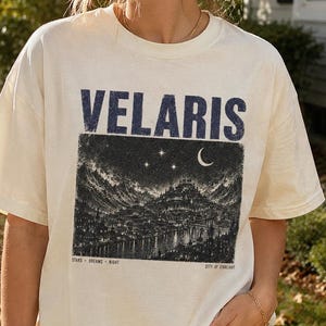 Puede incluir: Una camiseta de manga corta color hueso con un estampado gráfico azul oscuro. El texto VELARIS aparece sobre una ilustración detallada en blanco y negro de un paisaje urbano bajo un cielo estrellado con una luna creciente y el texto STARS DREAMS NIGHT CITY OF STARLIGHT.