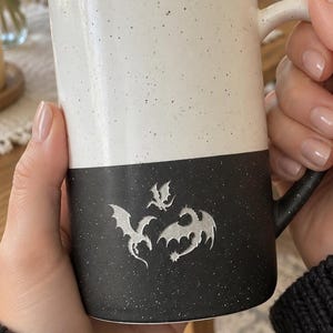 Może przedstawiać: Dwukolorowy kubek ceramiczny z nakrapianym wykończeniem. Górna połowa jest biała, a dolna czarna. Na czarnej części znajduje się srebrzysta grafika przedstawiająca trzy małe smoki w locie. Kubek posiada zakrzywione ucho po prawej stronie.