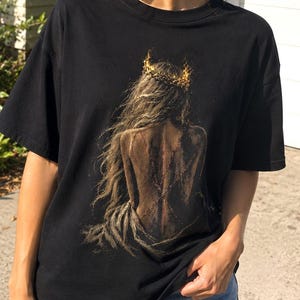 May include: A black short-sleeved t-shirt featuring a graphic print on the front. The design depicts the back view of a person with long hair wearing a crown made of flames. The artwork uses dark, earthy tones with bright orange and yellow highlights.
