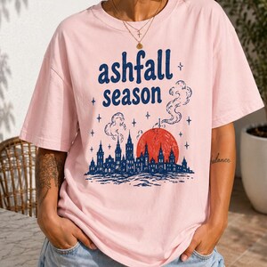 Puede incluir: Camiseta rosa claro con la frase "ashfall season" en azul, sobre un gráfico de una silueta de ciudad, un sol rojo y humo. La camiseta se combina con pantalones cortos de mezclilla.